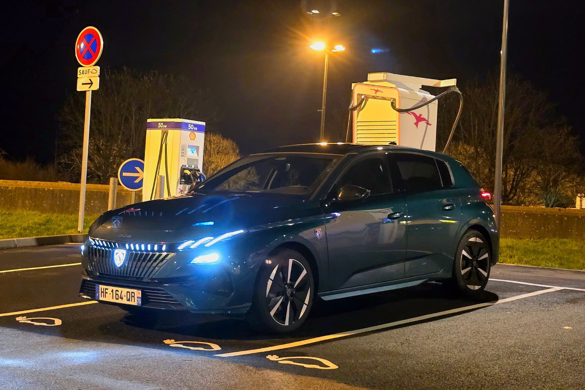 Nouvelle Peugeot 308 électrique 2026 de couleur Bleu Lagoa en cours de recharge nocturne sur une borne Ionity avec logo rétroéclairé visible
